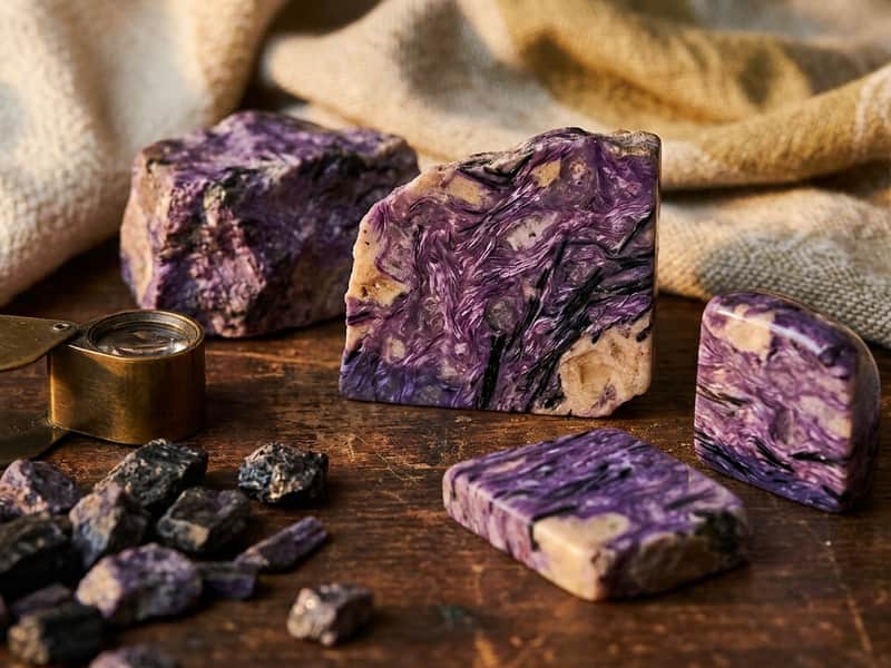 Raw and polished charoite specimens showing swirling lilac-purple patterns with black needle-like inclusions, beside a brass geologist's loupe on dark wood