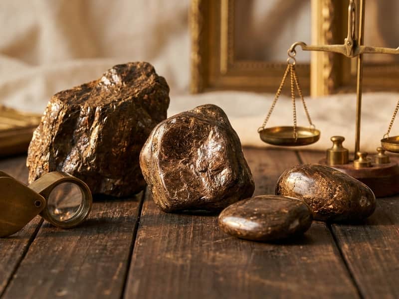 Raw and polished bronzite mineral specimens showing their metallic bronze-brown sheen, arranged on dark wood beside a brass jeweler's loupe and antique balance scale